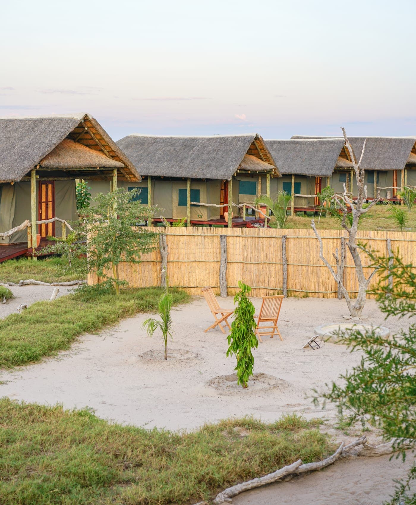Thatched tented rooms in a row at dusk, each with its own veranda and deck chairs on the sand