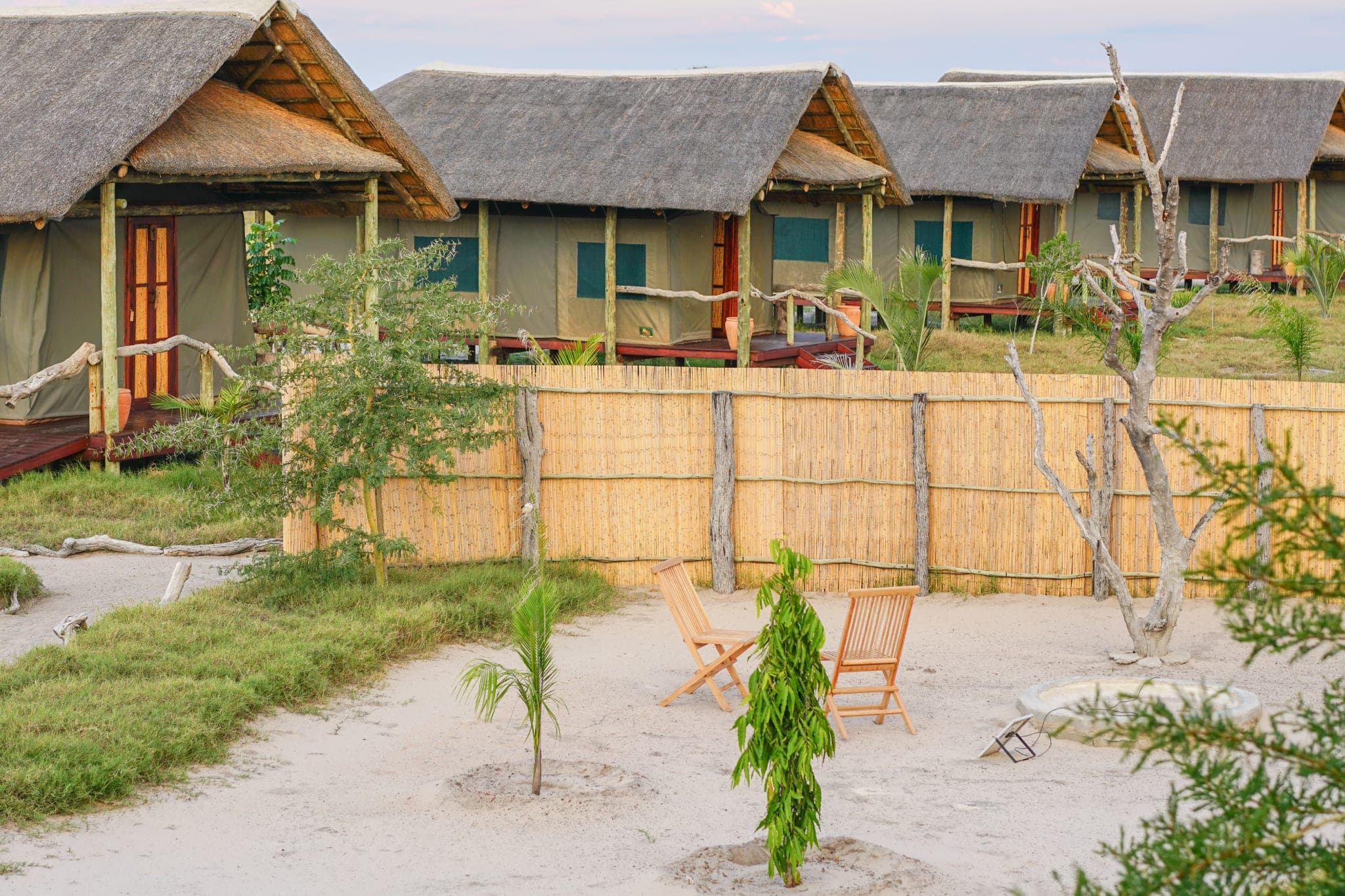 Thatched tented rooms in a row at dusk, each with its own veranda and deck chairs on the sand